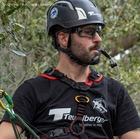 Man wearing upMOTION SRT climbing harness, black helmet and goggles, holding a drinking tube in his mouth, trees and leaves in the background