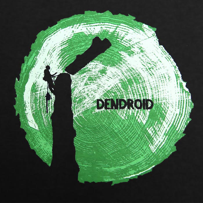 Close-up of green tree trunk cross-section with black tree climber and black text "DENDROID" on the black t-shirt "Go Big or Go Home", manufacturer not visible