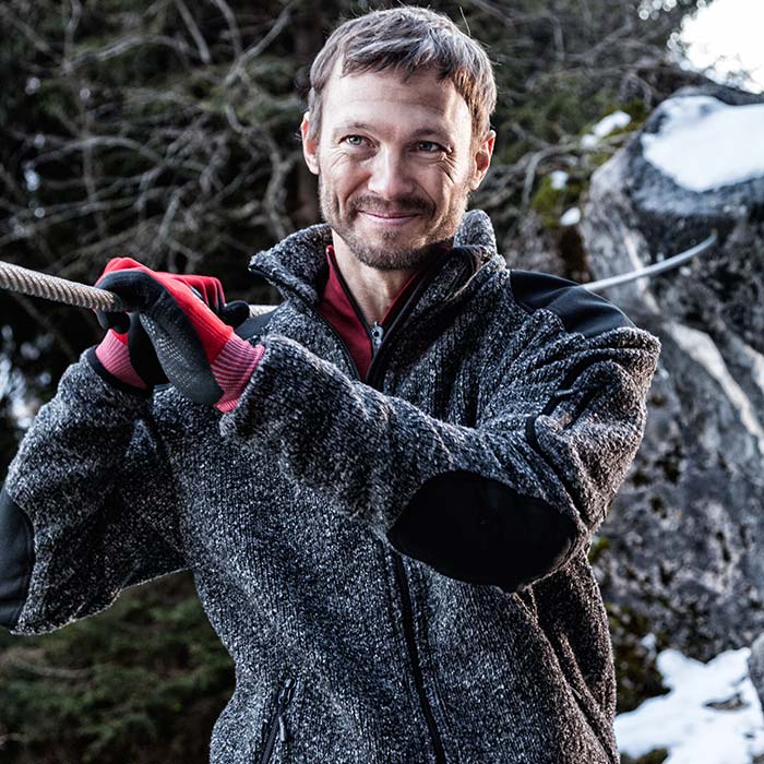 Man wearing grizzly jacket gray cardigan with black trim on shoulders and elbows, red undershirt and red gloves, forest and rocks with snow in the background