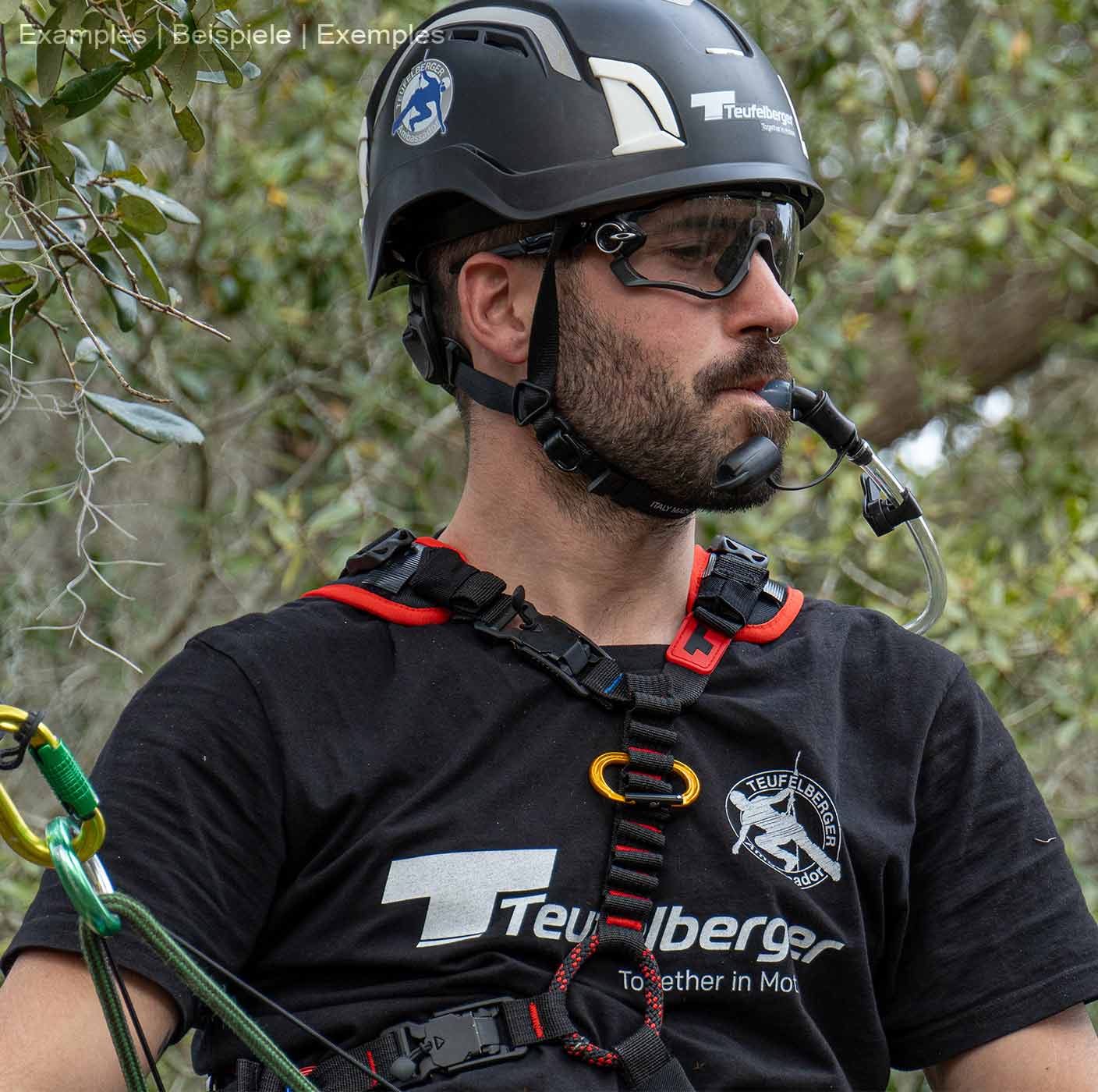 Man wearing upMOTION SRT climbing harness, black helmet and goggles, holding a drinking tube in his mouth, trees and leaves in the background