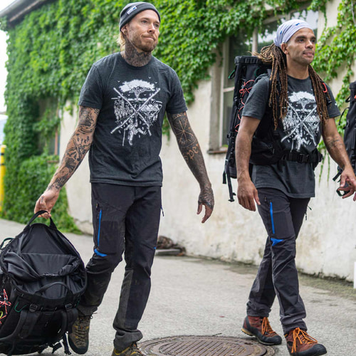 Two men are wearing the dark grey tSPIRIT T-shirt with a white climbing and nature motif, one is wearing a black cap and holding a bag, the other is wearing a light-coloured headscarf and a backpack