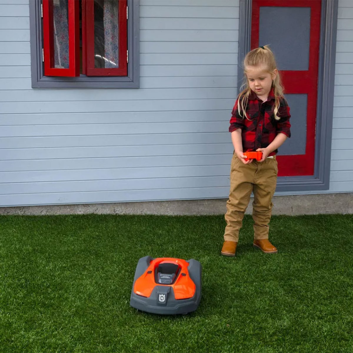 A child in a red checked shirt, beige pants and brown boots controls the orange and black children's Automower from Husqvarna on a green lawn in front of a house with a red window and door frame