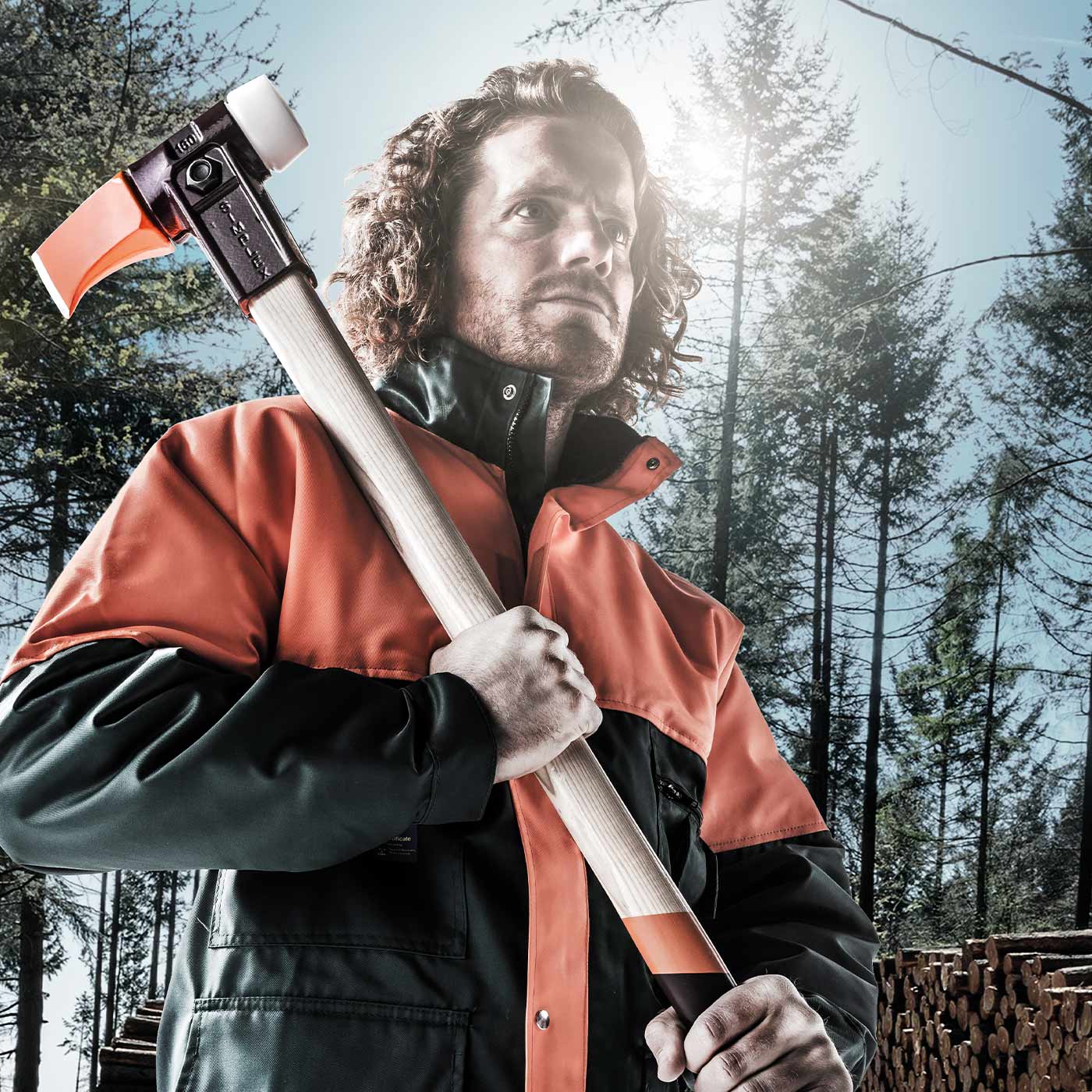 Man in orange and black work suit holding the Simplex 900 axe on his shoulder, forest and stacked tree trunks in the background