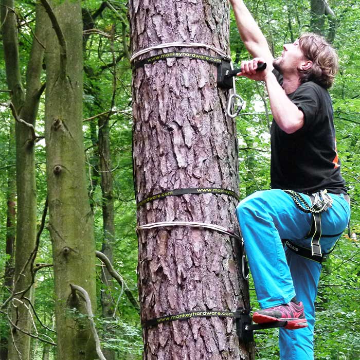 Tree-Monkey Steppers attached to tree trunk, man with blue trouser leg and black t-shirt climbs with foot on metal foot and harness