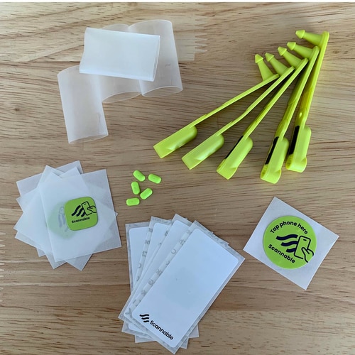 Climber Pack NFC Tag with yellow plastic parts, several white sleeves, green stickers with Scannable logo and small yellow components on a wooden table