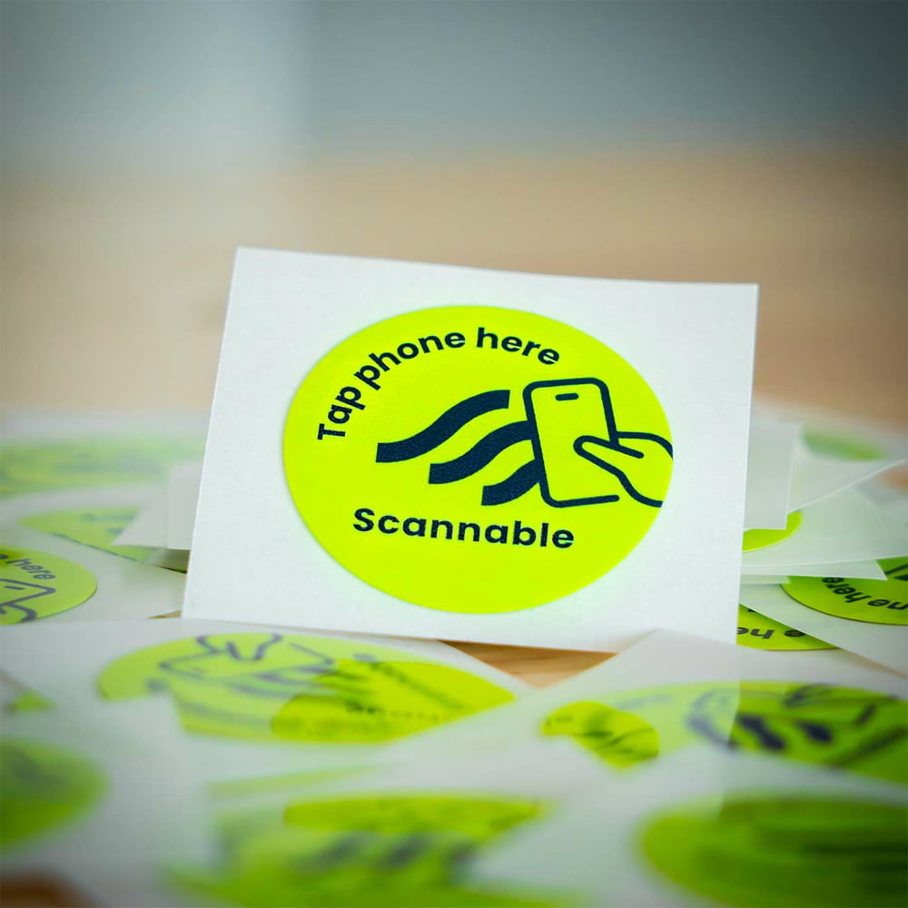 Close-up of a single round, yellow helmet sticker NFC tag sticker with black inscription "Tap phone here Scannable" and a smartphone icon, surrounded by more stickers
