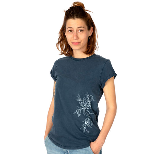 A woman wears a dark blue T-shirt "Olive Branch" with a white olive branch motif on the side and has her hands in her trouser pockets.