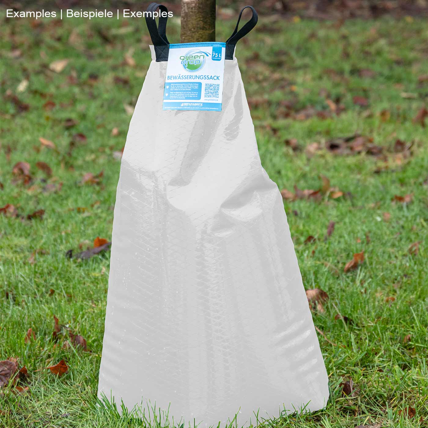 Greendrop White Watering Bag in White stands on green grass, with two black carrying loops and blue label at the top center, surrounded by autumnal leaves on the ground