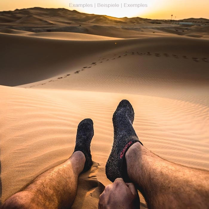 Skinner's black sock with rubberized sole on one foot in the desert, in the background sand dunes at sunset