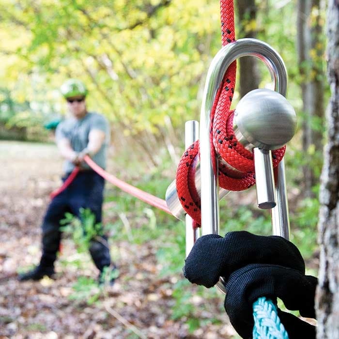 Port A Wrap Large made of shiny metal to which red and blue ropes are attached, in the forest, with a person in the background holding a rope.