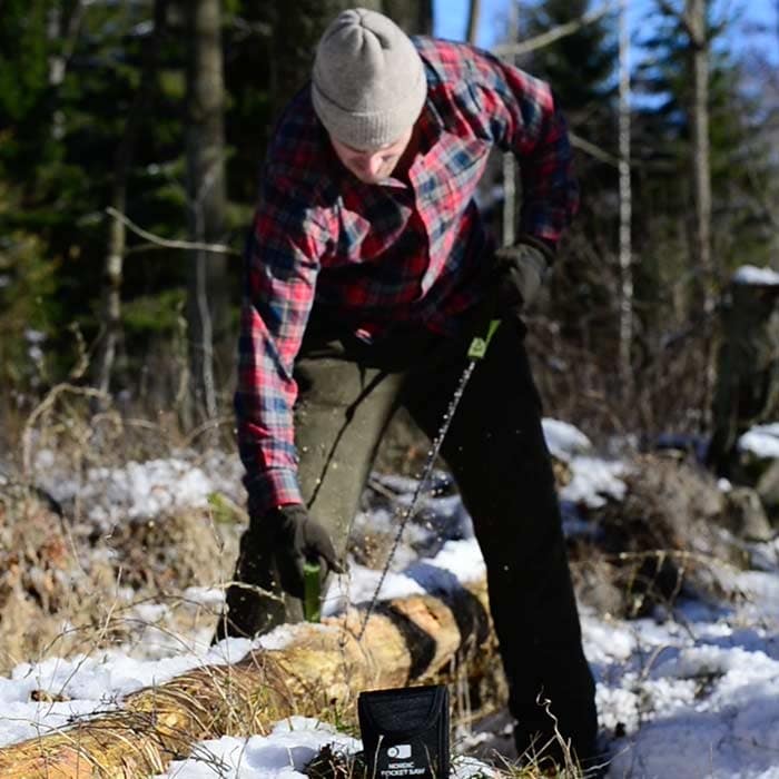 Person with gray cap and checkered shirt using the Nordic Pocket Saw on a tree trunk, black bag with white lettering lying on the floor