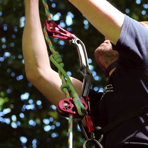 Person in black shirt holding Rope Wrench Squirrel PPE Regulations System Kit with red and black metal device, green and white rope and black carabiner from ISIC, background green leaves and sky