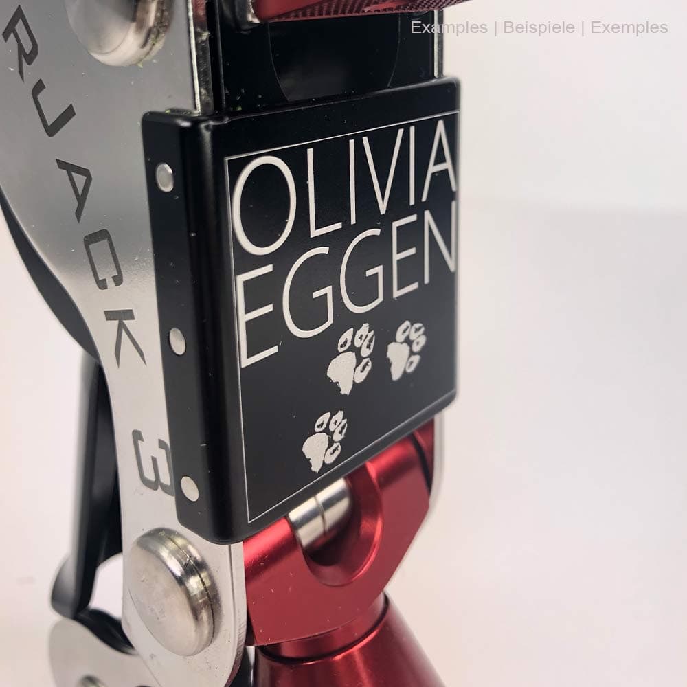 Close-up of the black shield on the SpiderJack 3 with white engraving "OLIVIA EGGEN" and paw prints, red metal hook and silver frame
