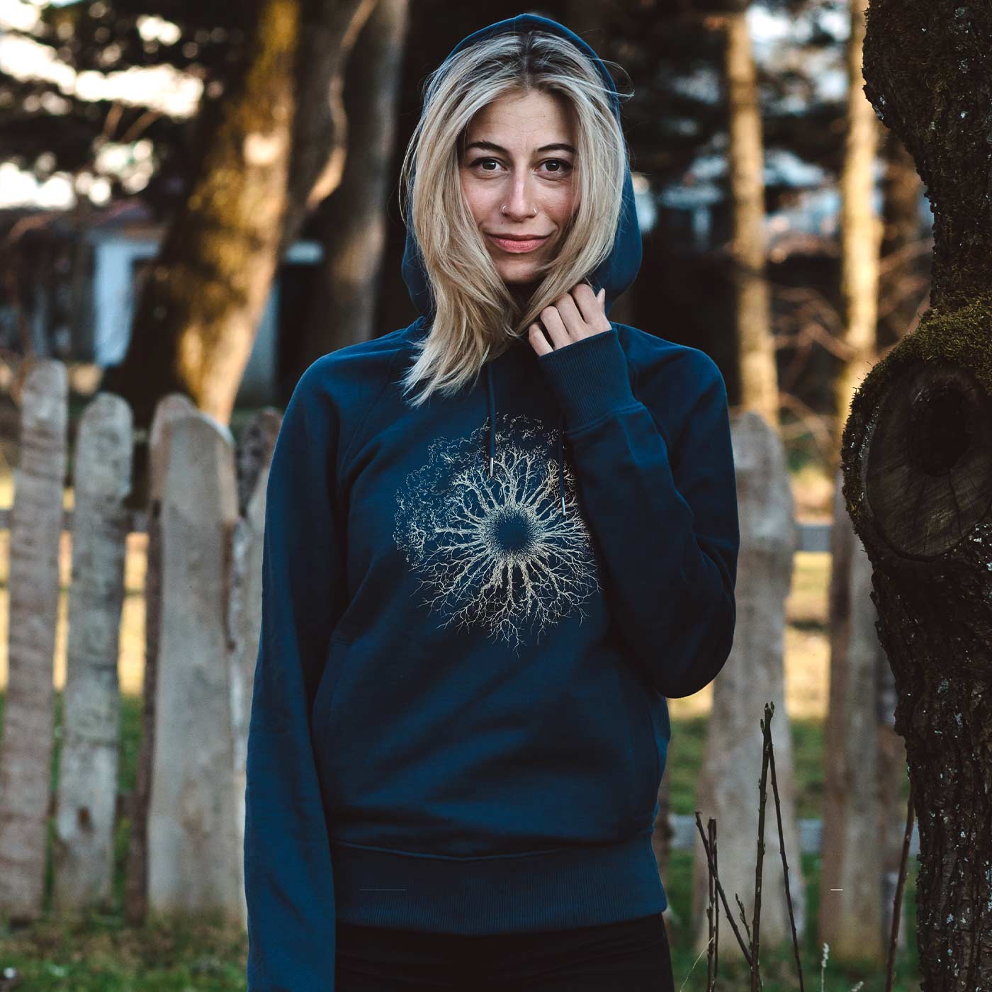 A woman stands outside next to a tree, wearing a blue Wooden Iris hoodie with a hood and a white, treeroot-like pattern on the chest.