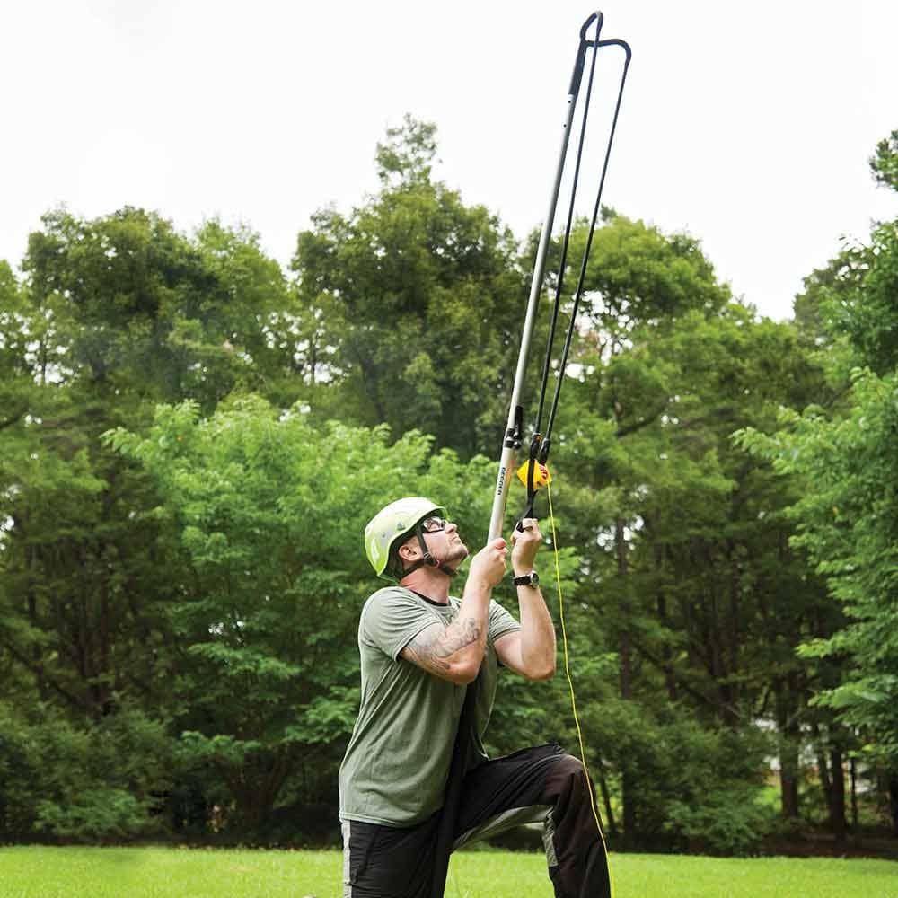 Person in green helmet and goggles kneeling on green grass and holding Big Shot wand with black handle and yellow webbing in front of a forest