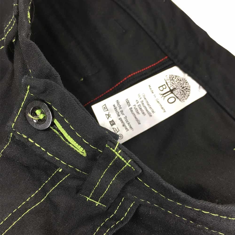 Close-up of the waistband of the Roots Pure outdoor pants in black with green buttonhole and green thread, inside a white label with text and tree logo