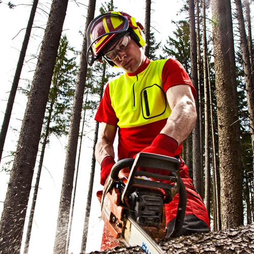 Man wearing the zip-neck shirt in red and yellow, safety helmet, goggles and gloves, holding a chainsaw in the forest