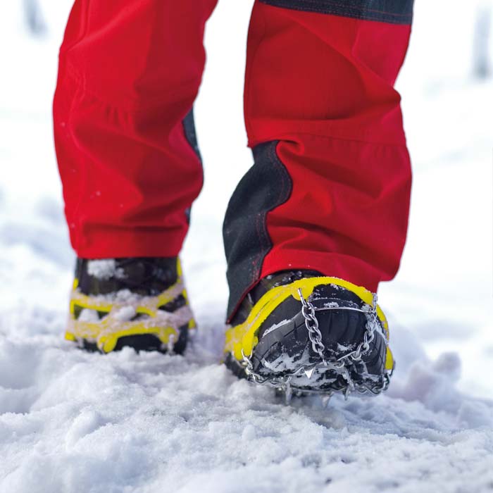 Spikes with yellow elastic bands and metal chains on black shoes, worn by a person in red pants in the snow