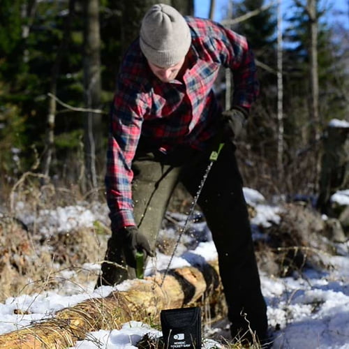 Person with gray cap and checkered shirt using the Nordic Pocket Saw on a tree trunk, black bag with white lettering lying on the floor