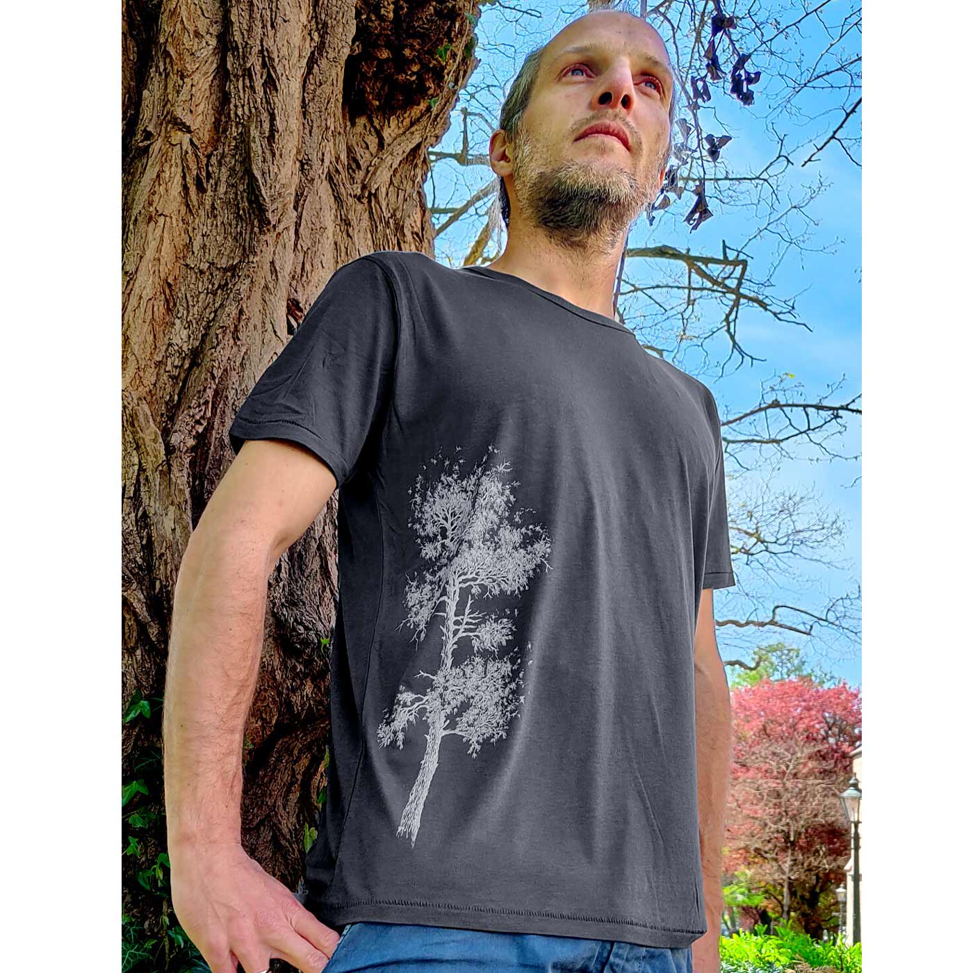 Man standing outside in front of a tree wearing a dark gray Pine Tree T-shirt with a white tree print on the left side of the shirt.