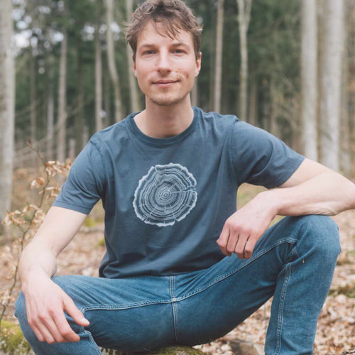 A man sits in the forest and wears a dark blue T-shirt with a white tree trunk cut, product name Treeslice