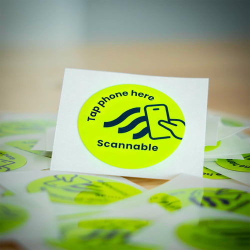 Close-up of a single round, yellow helmet sticker NFC tag sticker with black inscription "Tap phone here Scannable" and a smartphone icon, surrounded by more stickers