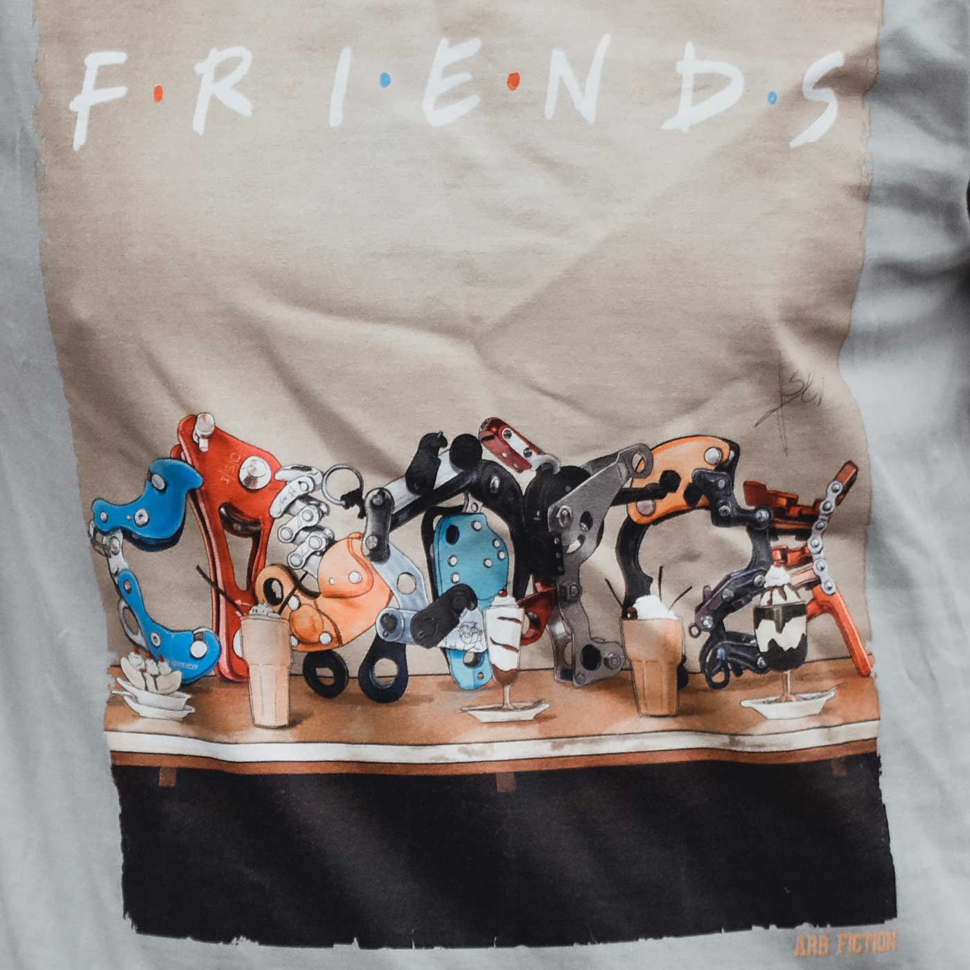 Close-up of the T-shirt print with the words "Friends" and detailed mechanical parts in different colors on a table, with the lettering "ARB FICTION" visible at the bottom right