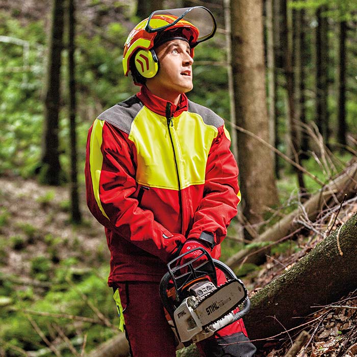 Man wearing cut-resistant jacket in red with yellow and gray accents, helmet and gloves, holding a chainsaw in the forest