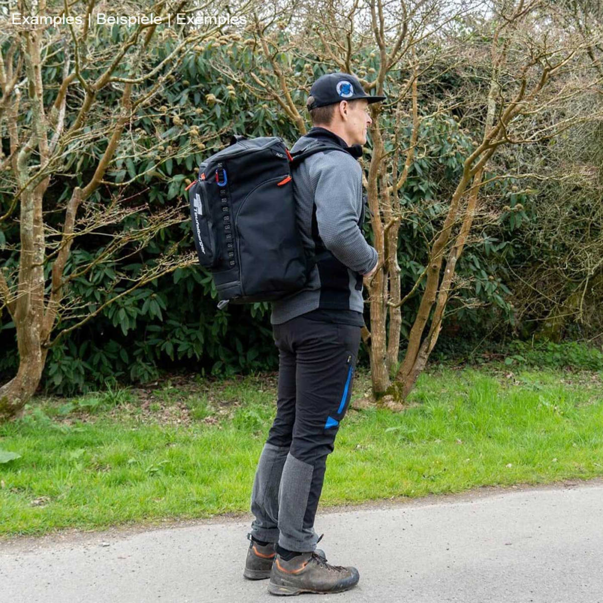 Person with black backpack baby mule, manufacturer not visible, standing sideways on a path, surrounded by trees and grass