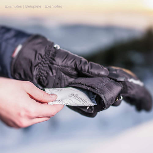 Hand warmer from thermopad is held in the hand by a person in black winter glove half, the heat cushion is printed white with black thermopad lettering