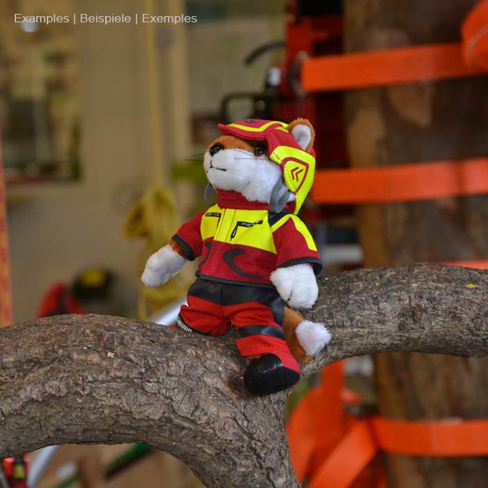 Wiffzack plush figure sitting on a tree trunk, wearing red and yellow clothes and helmet, in the background the workshop environment visible