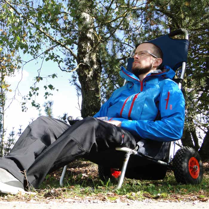 Man sitting in a dark blue beach rolly beach wheelchair outdoors surrounded by trees