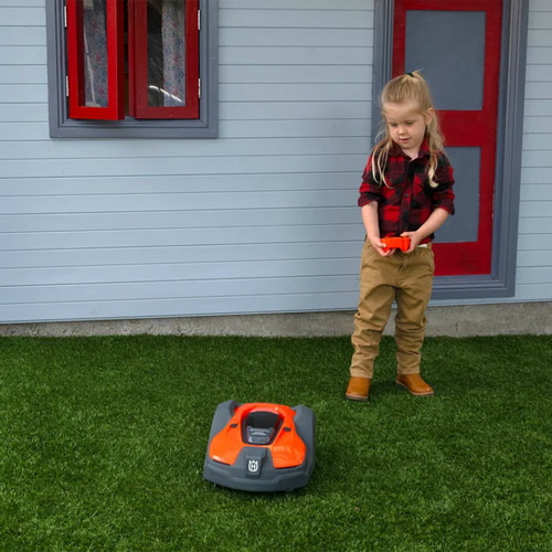 A child in a red checked shirt, beige pants and brown boots controls the orange and black children's Automower from Husqvarna on a green lawn in front of a house with a red window and door frame