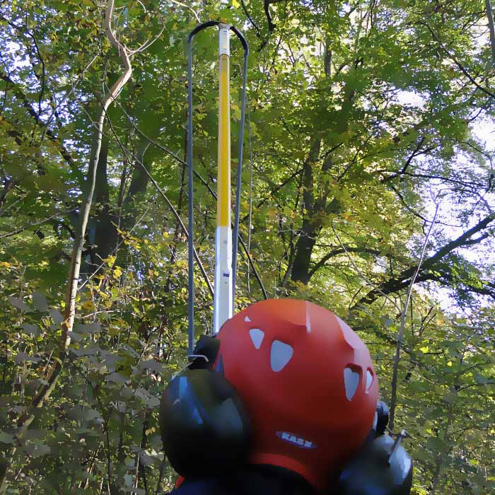 Person with red helmet and ear protection from behind, holding the Big Shot rod with black handle and yellow webbing in a forest