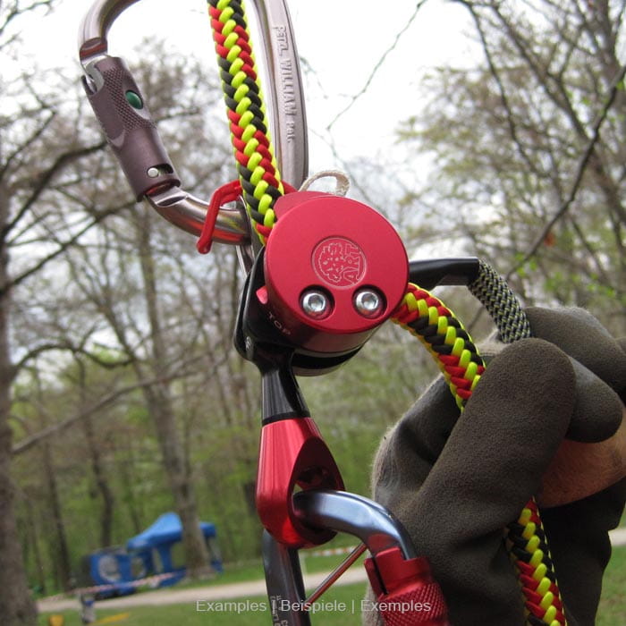 LockJack 2 climbing frame with red metal housing, grey carabiner and yellow-red rope, held by a hand with grey glove against a forest background