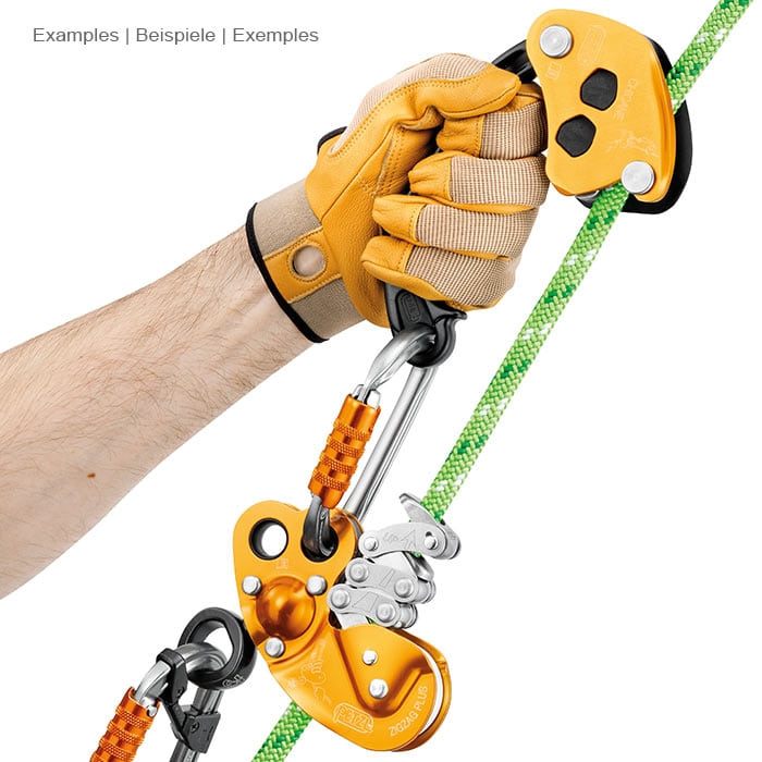 Petzl Chicane, yellow and black belay device with handle, held by a hand with yellow glove, connected to a carabiner and a yellow belay device on a green rope