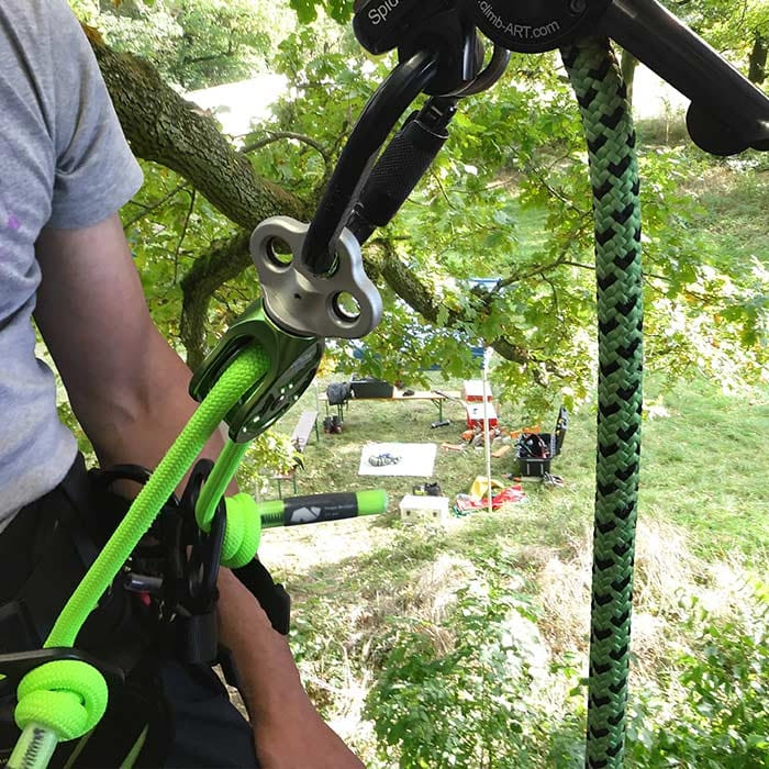 Person wearing gray t-shirt carrying a green and silver rook pulley attached to a black snap hook with rope, trees and grass in the background
